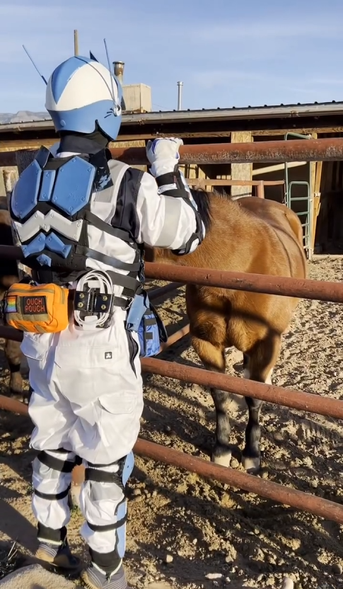 Person dressed in a futuristic blue and white costume with a helmet, backpack, and orange pouch, standing near a brown horse in a fenced outdoor area.