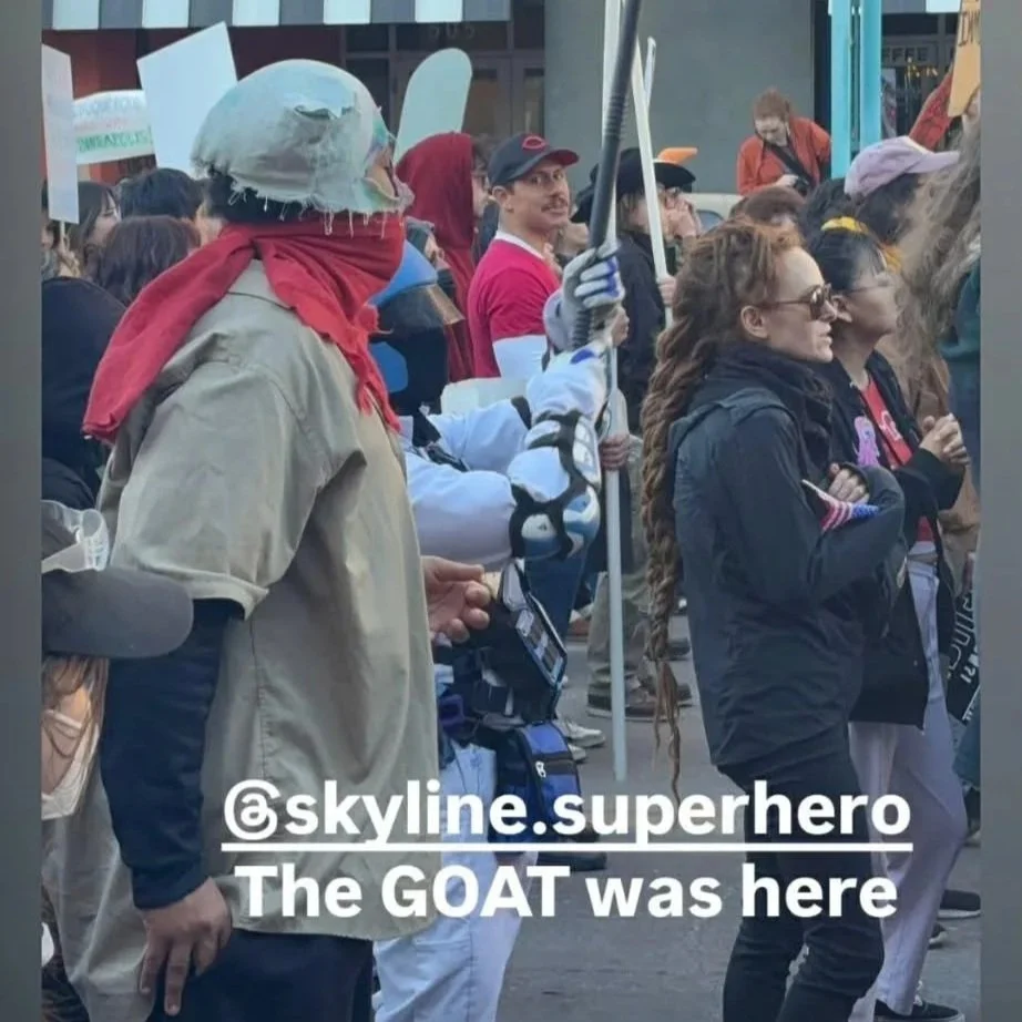 People dressed in cosplay costumes participating in a parade or event, with a person wearing a makeshift goat mask and red scarf in the foreground.