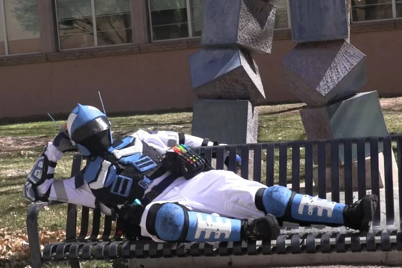 A person dressed in a futuristic robot costume lying on a park bench with their head resting on one hand and legs crossed, next to modern abstract stone sculptures.