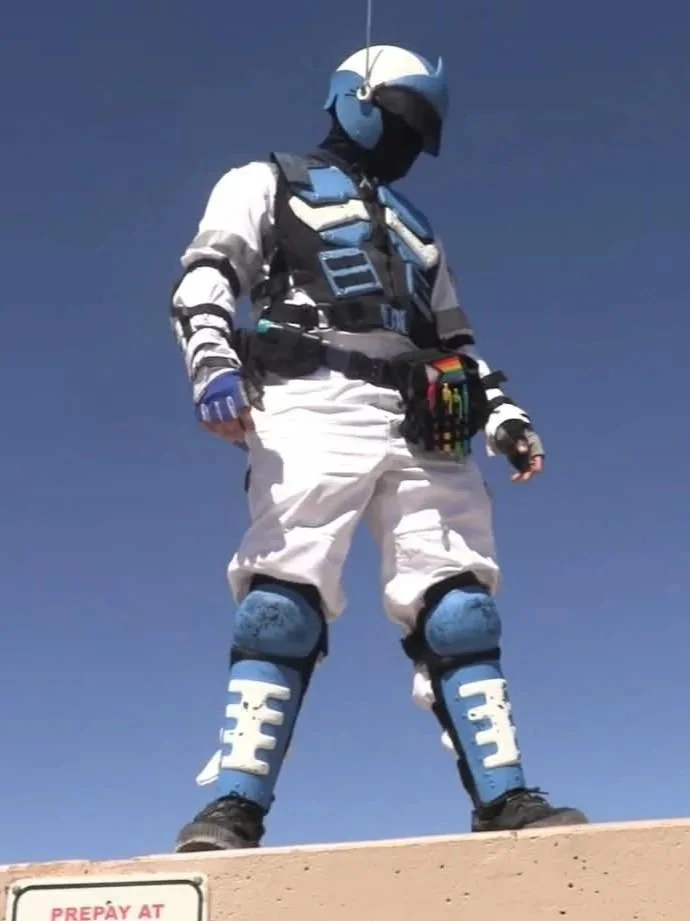 A person standing outdoors dressed in with astronaut-themed gear, including a helmet, padded suit, and knee protectors, with a parachute harness visible, against a clear blue sky.