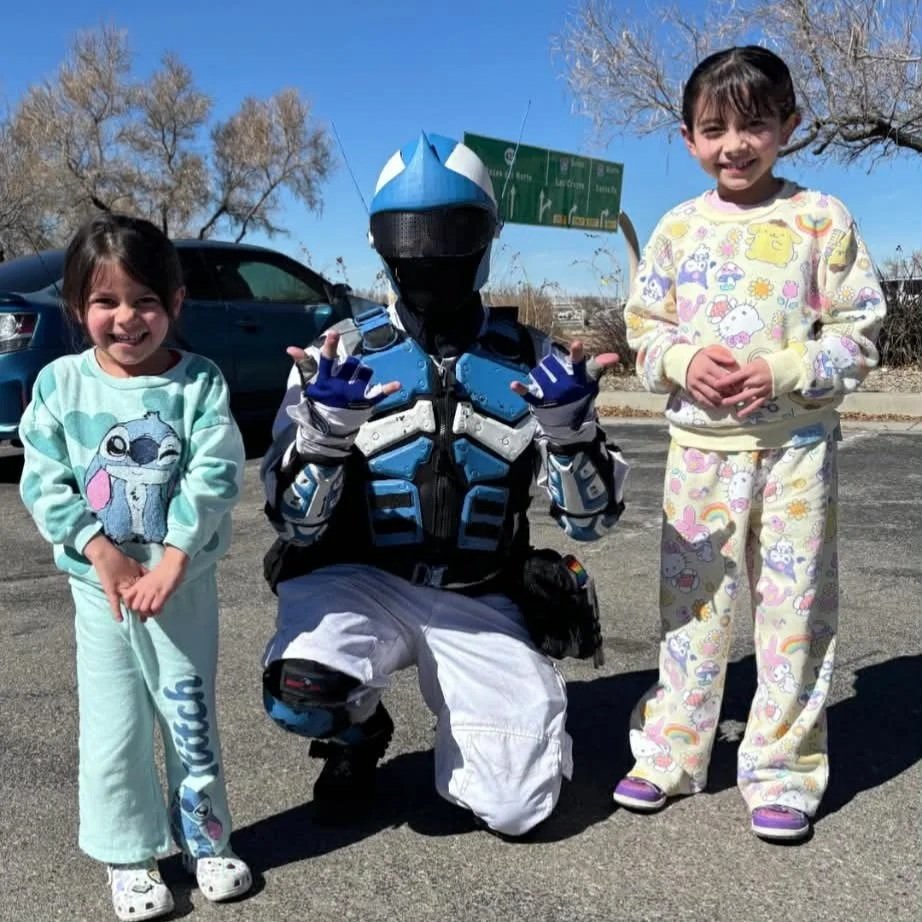 Two young girls in pajamas and a person in a detailed blue costume of a character from a video game, all standing outside on a parking lot on a sunny day.
