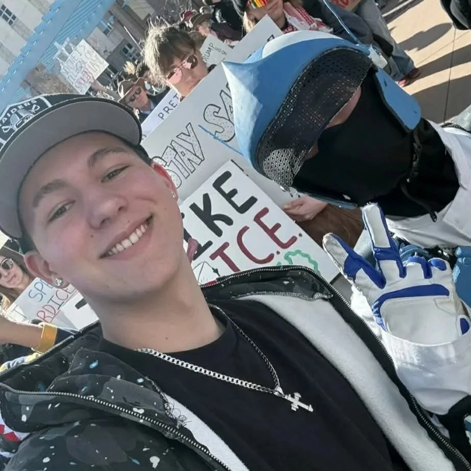 Young man smiling at a protest or rally, wearing a baseball cap, black hoodie, and necklace with a cross, holding up a peace sign with a gloves. Other protesters and signs are visible in the background.