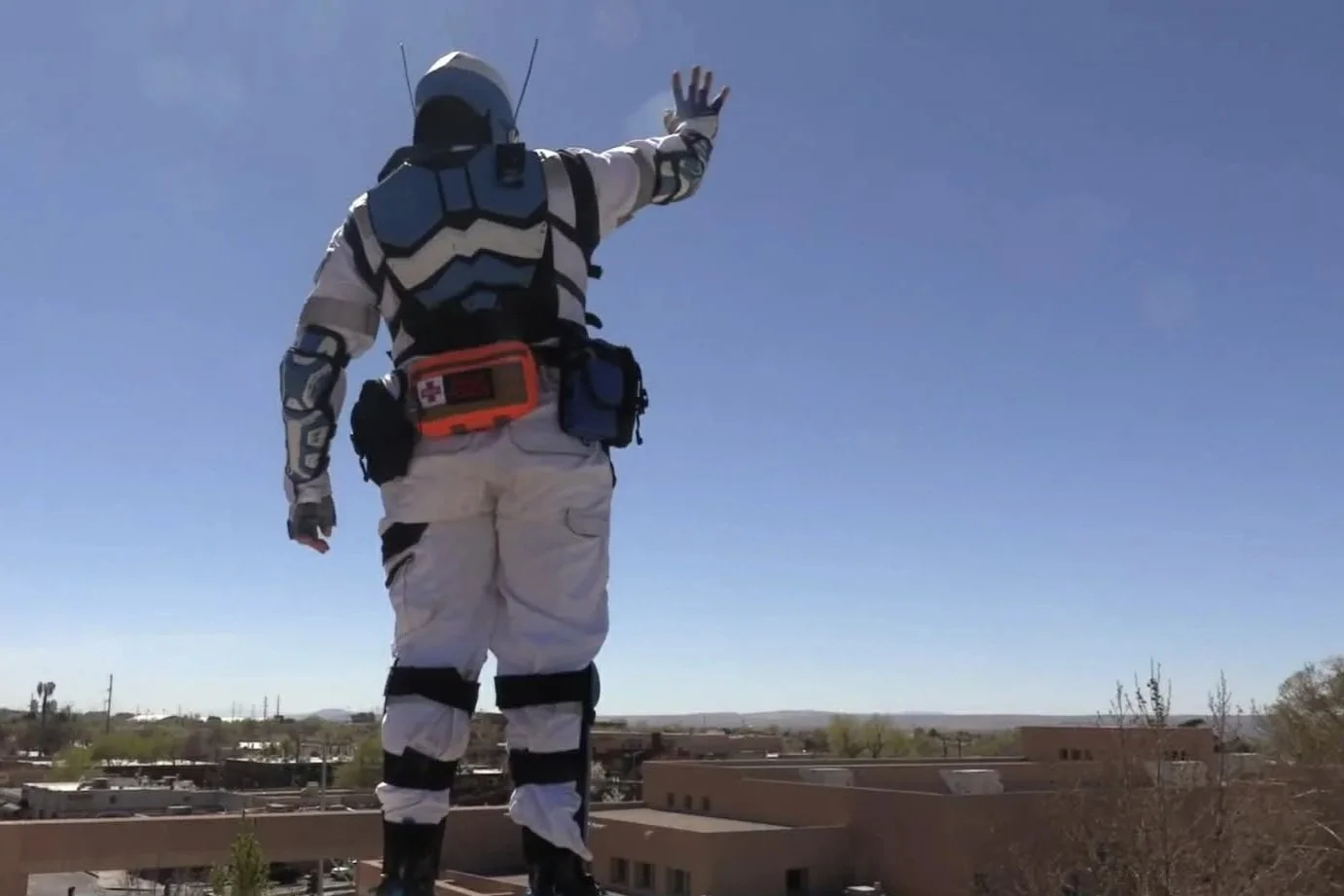 Astronaut in a space suit standing on a rooftop, waving to the sky on a clear day with buildings and trees in the background.