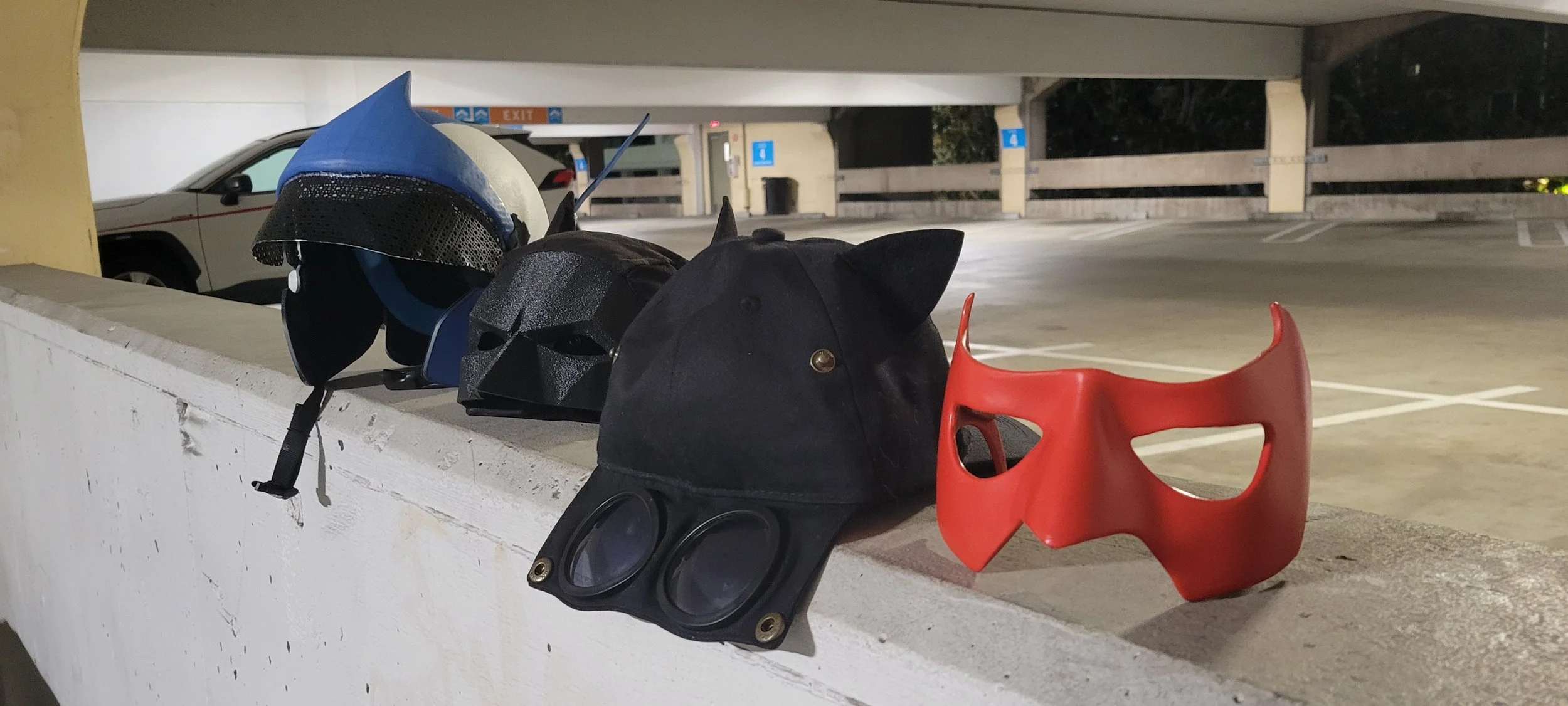 Various masks and helmets, including a blue helmet, a black Darth Vader helmet, a black cap with animal ears, and a red superhero mask, placed on a ledge in a parking garage.
