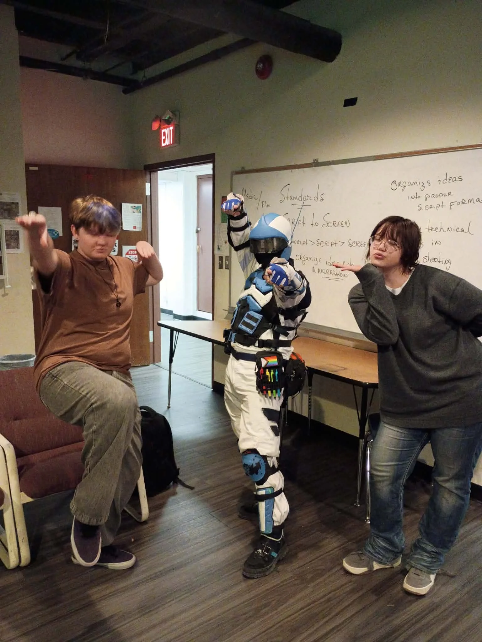 Three people in a room posing with playful gestures near a whiteboard filled with notes, a person in a striped costume and mask holding a shield, and a young man with short brown hair and glasses.