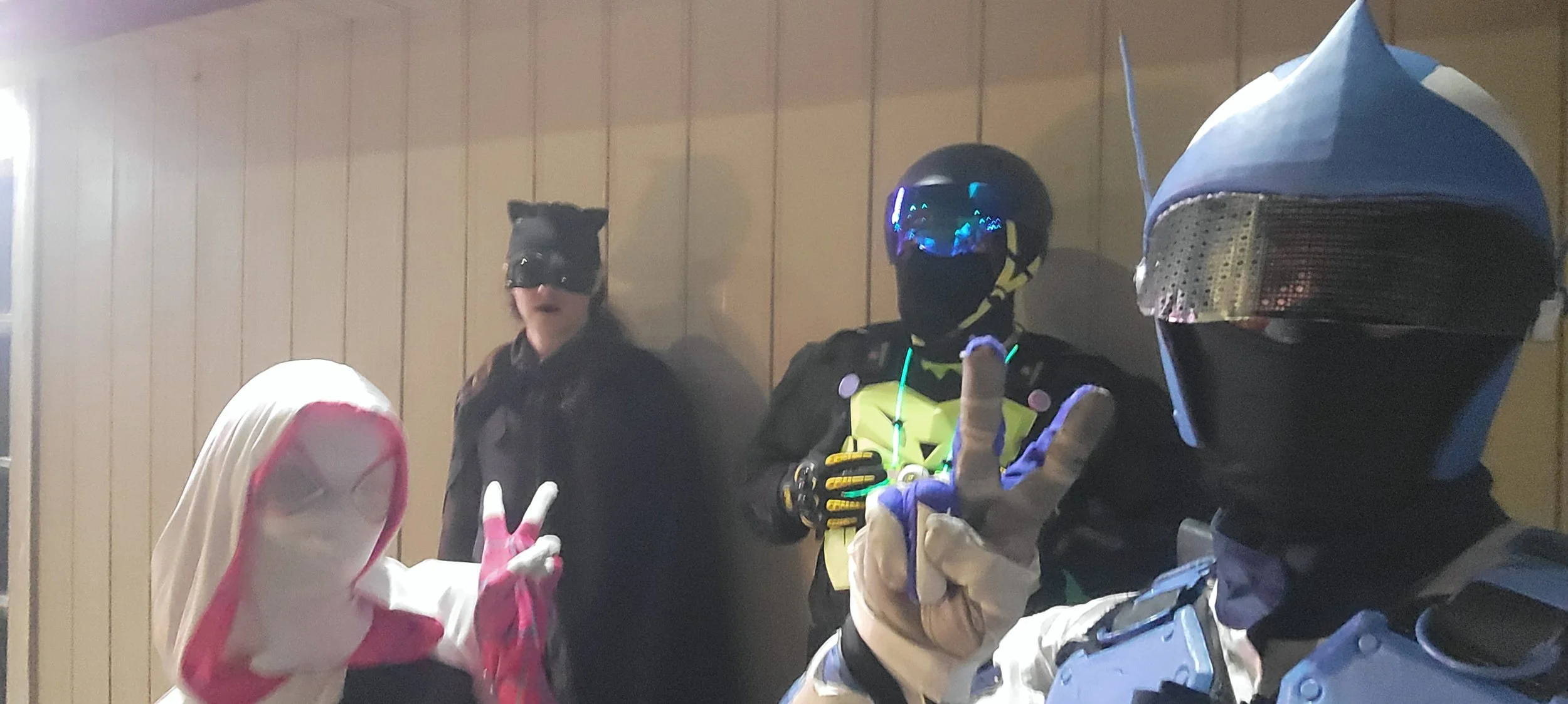 Three people in costume and gear posing indoors, one wearing a black mask and helmet with a reflective visor, another in a black and yellow suit with a helmet and reflective visor, making a peace sign, and the third in a pink and white hooded outfit 