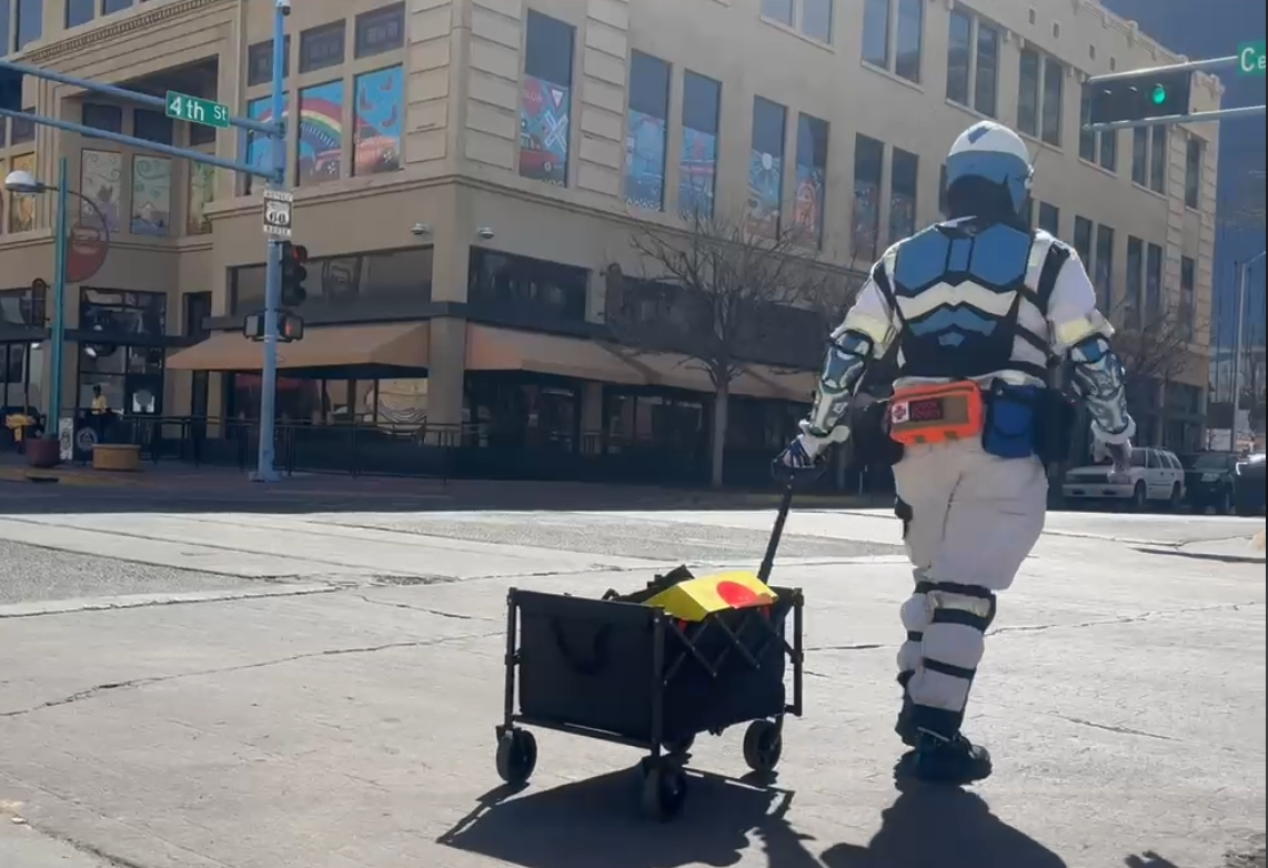 A person in astronaut costume pulling a black utility cart on a city street during daytime, with a multi-story building and traffic lights in the background.