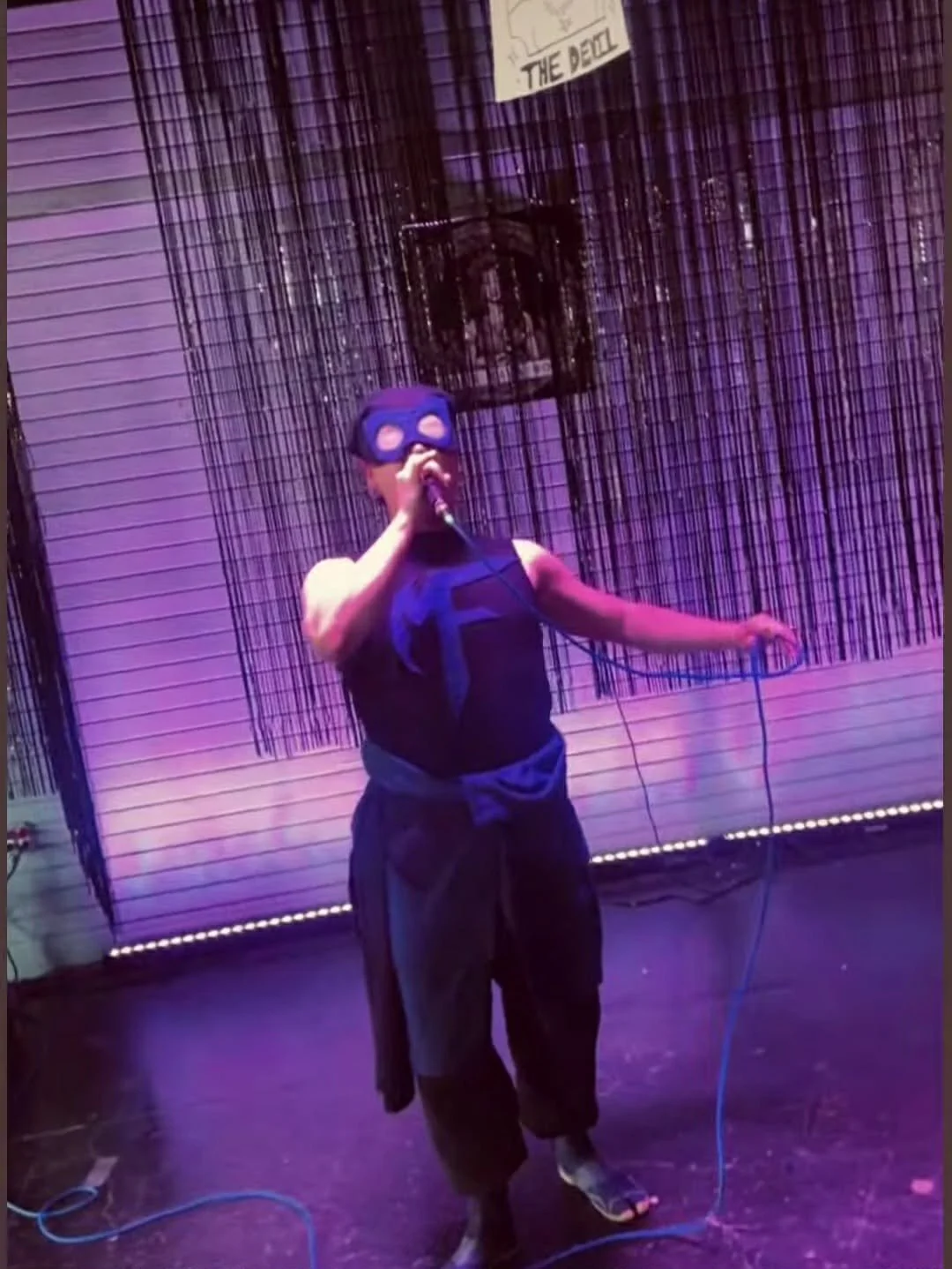 Person wearing a ski mask singing into a microphone on a stage with purple lighting and a decorative backdrop.