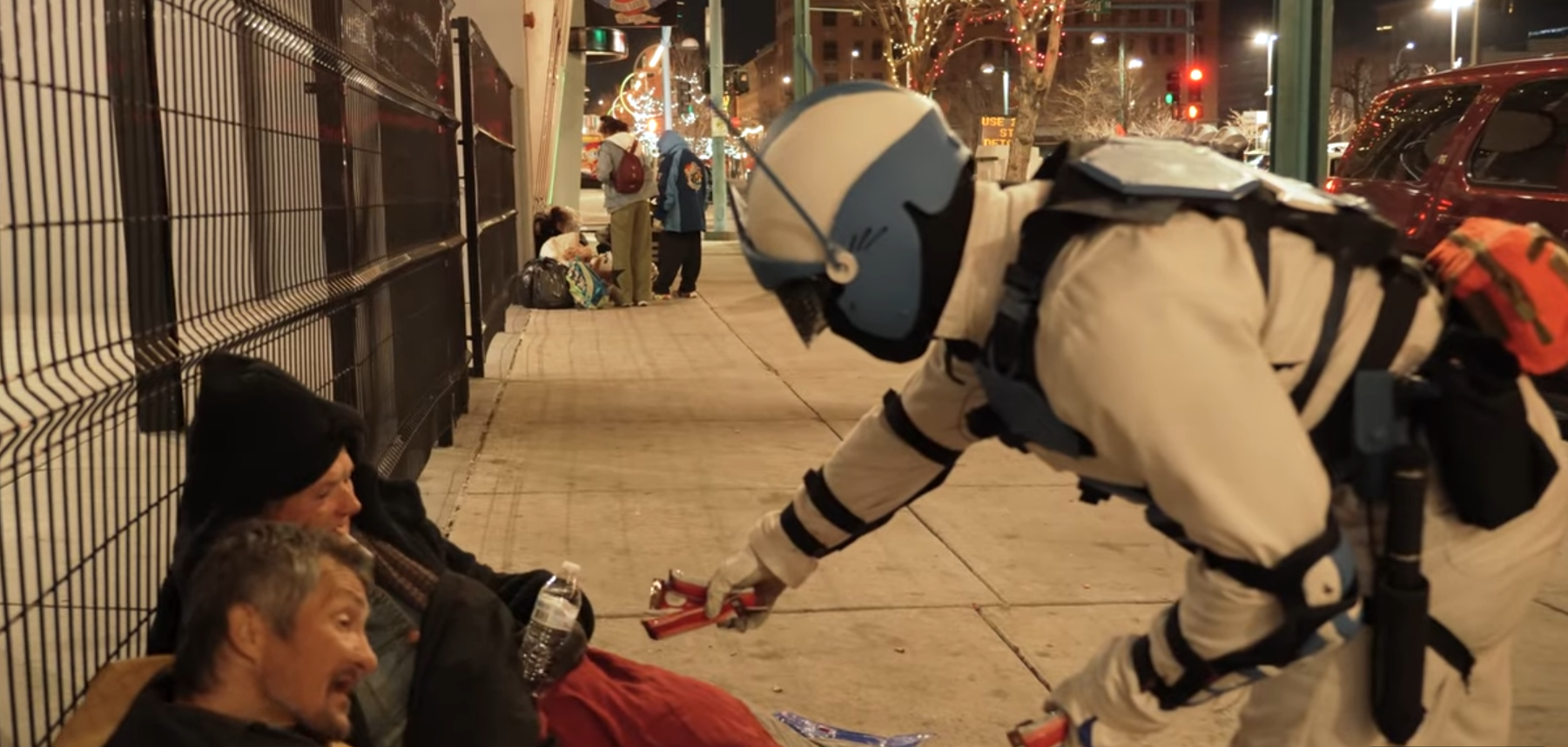 A person in a white rescue uniform and helmet offers a water bottle and some supplies to two homeless individuals sitting on the sidewalk at night, with other homeless people and belongings visible in the background.