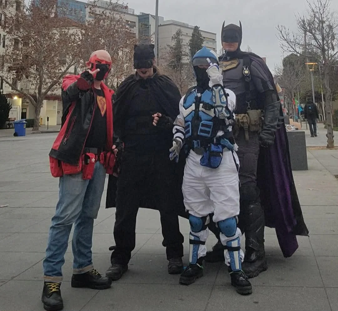 Four people dressed as superhero characters standing outdoors in an urban area with trees and buildings in the background.