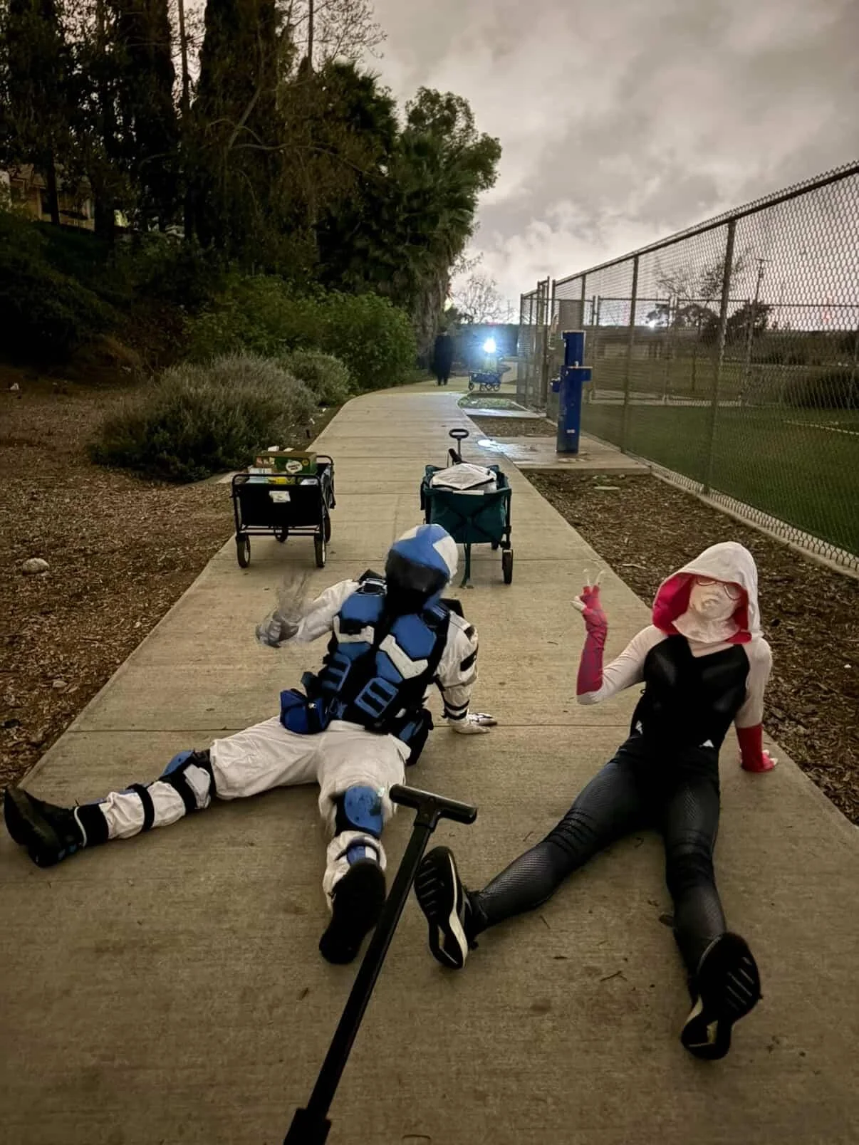 Two people dressed in superhero costumes sitting on a sidewalk near a fence and some trees. One dressed as a robot and the other as a person with a white hood and glasses. There are carts and personal belongings around them, with a light in the backg