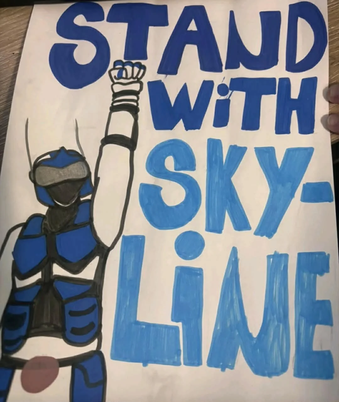 A hand-drawn sign with the message, 'STAND WITH SKY-LINE,' in large, bold blue letters. The sign features an illustration of a person in a blue and black striped sports outfit raising their fist, wearing a helmet with a visor and goggles, and giving 