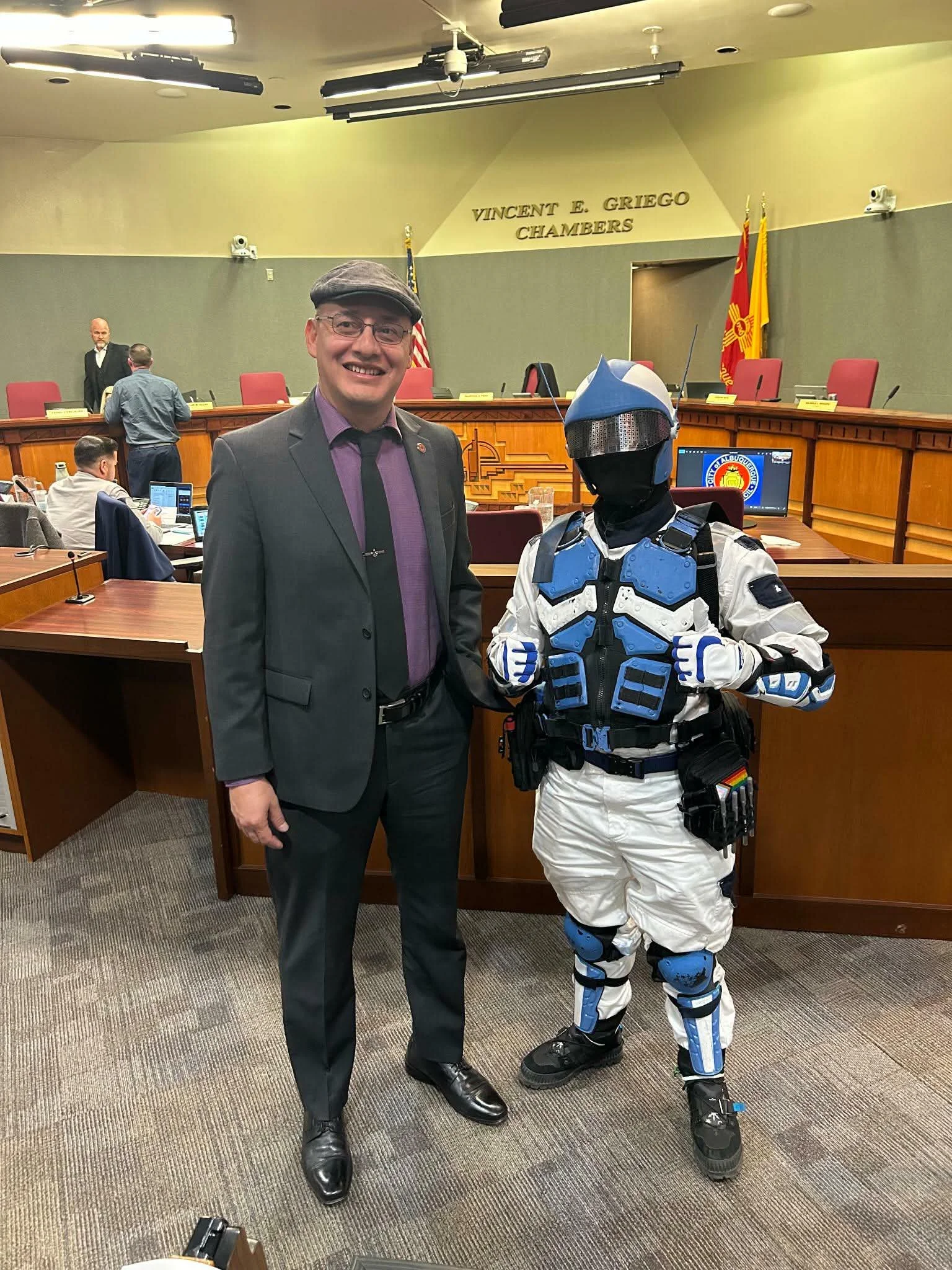 Photo of two men standing in a conference room, one in a suit and the other in futuristic armor with a helmet and gloves, with a sign reading 'VINCENT E. GREIGO CHAMBERS' in the background.