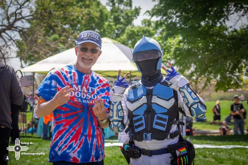 A man wearing a tie-dye shirt and a cap that says 'Air Force Veteran' stands next to a person dressed in a futuristic, blue and black costume with a helmet, making peace signs with their hands during an outdoor event with tents and trees in the backg