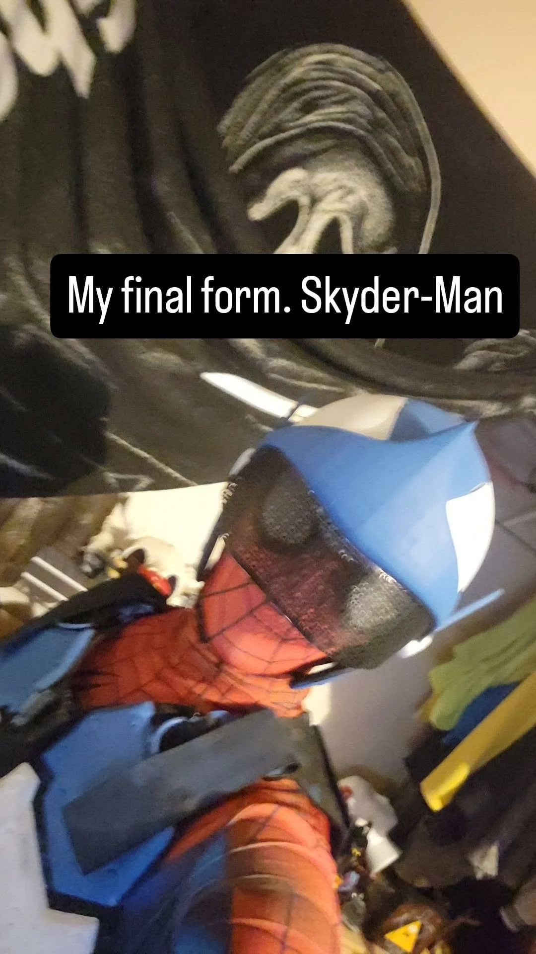 A person dressed in a Spider-Man costume taking a selfie in front of a black T-shirt printed with a white dragon and the words "My final form. Skyder-Man." A small dog lying on the floor in the background.