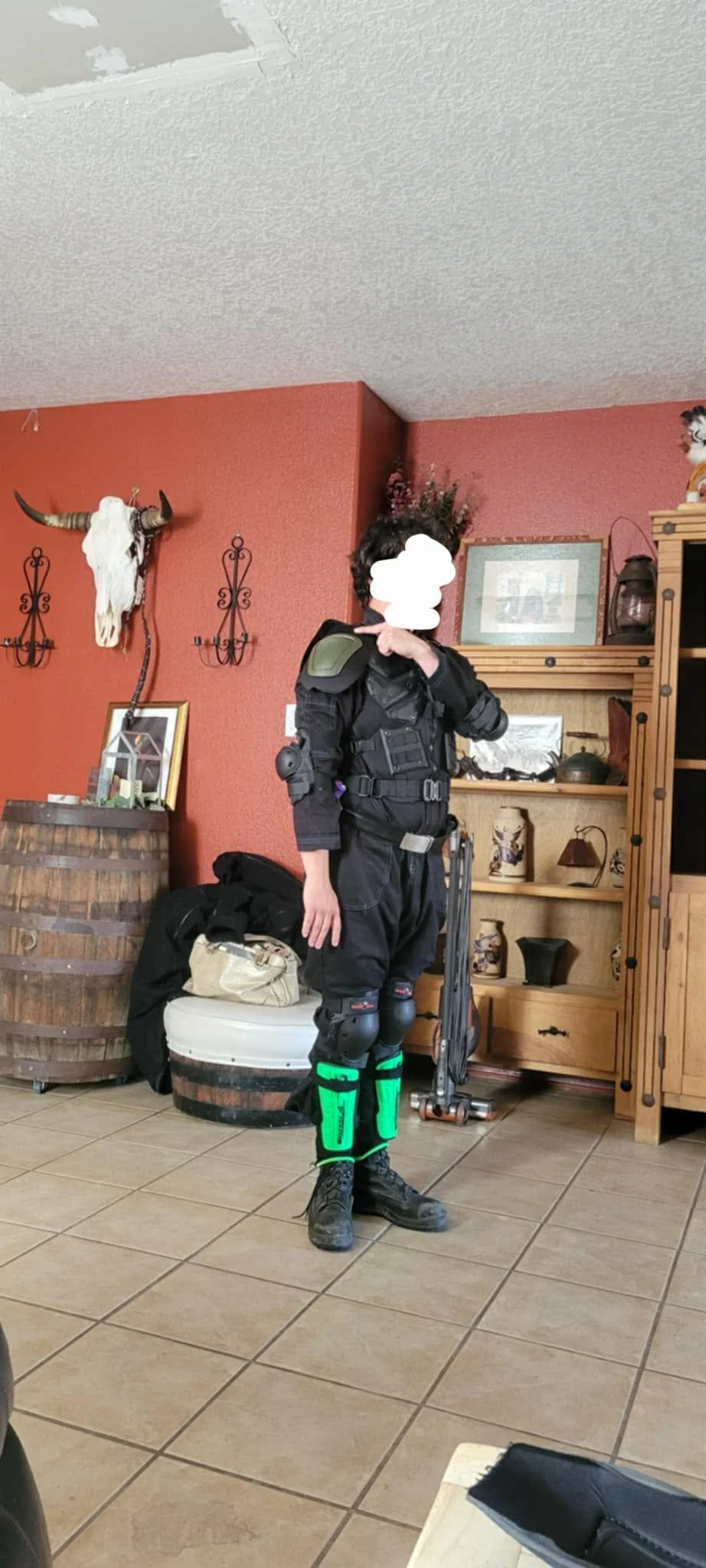 Person dressed in black tactical gear with knee and shin pads, standing indoors in front of a wooden shelving unit, with a red wall and decorative items including a mounted animal skull in the background.