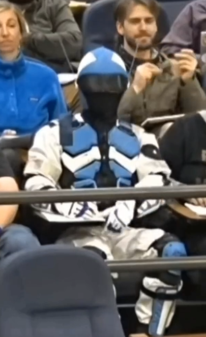 Person in a blue and white racing suit and helmet sitting among spectators at a sporting event.