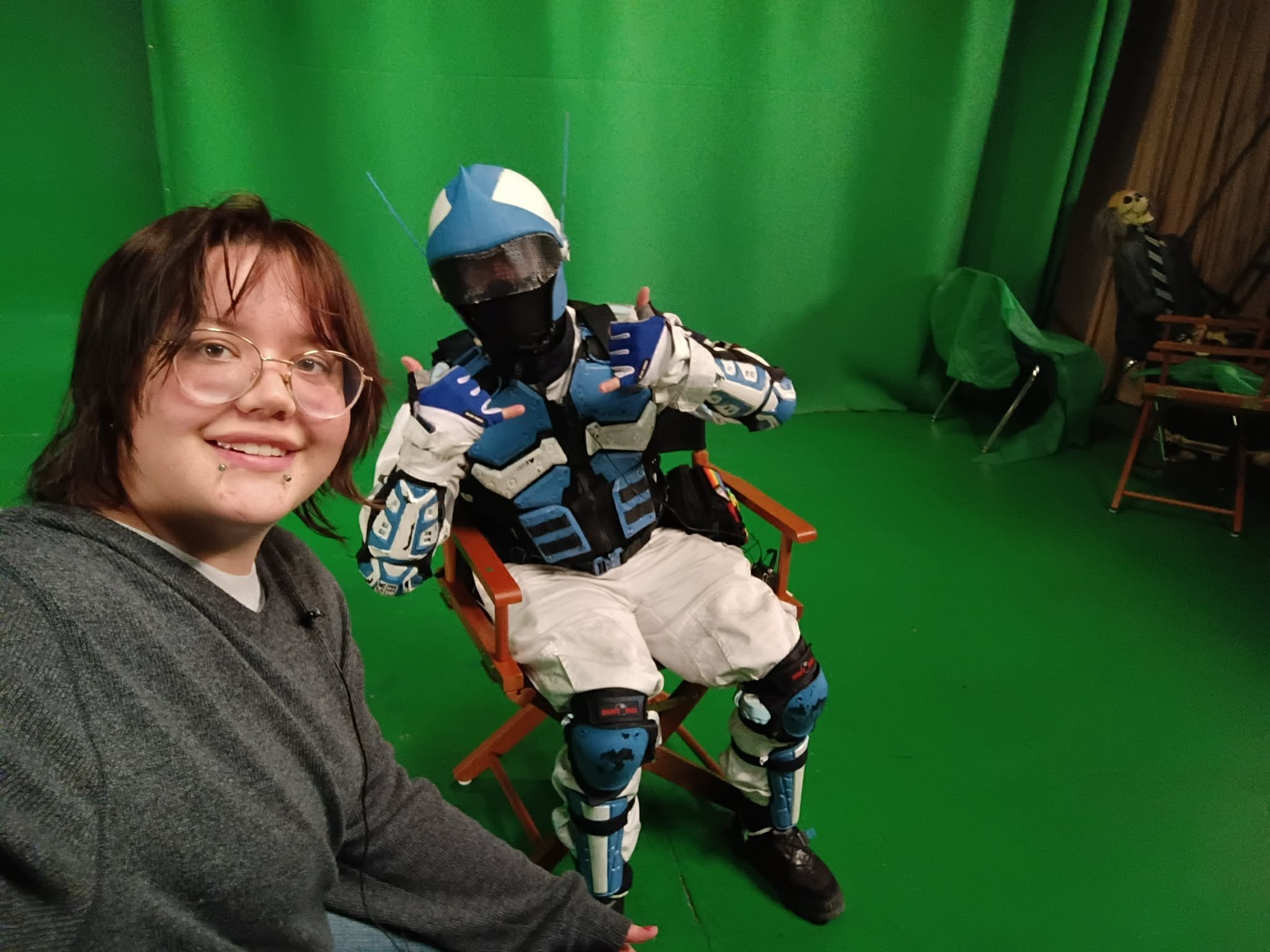 A woman with glasses and piercings taking a selfie with a person in a futuristic blue and black costume sitting on a director's chair in front of a green screen.