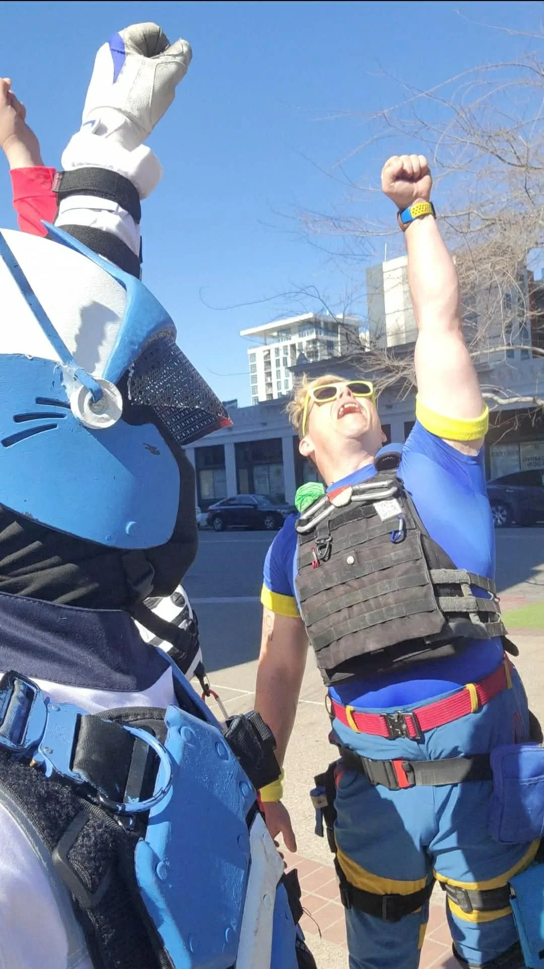 Two people outdoors, one wearing a motorcycle helmet and gear, and the other in a T-shirt with a vest and sunglasses, raising fists in a gesture of victory or celebration.