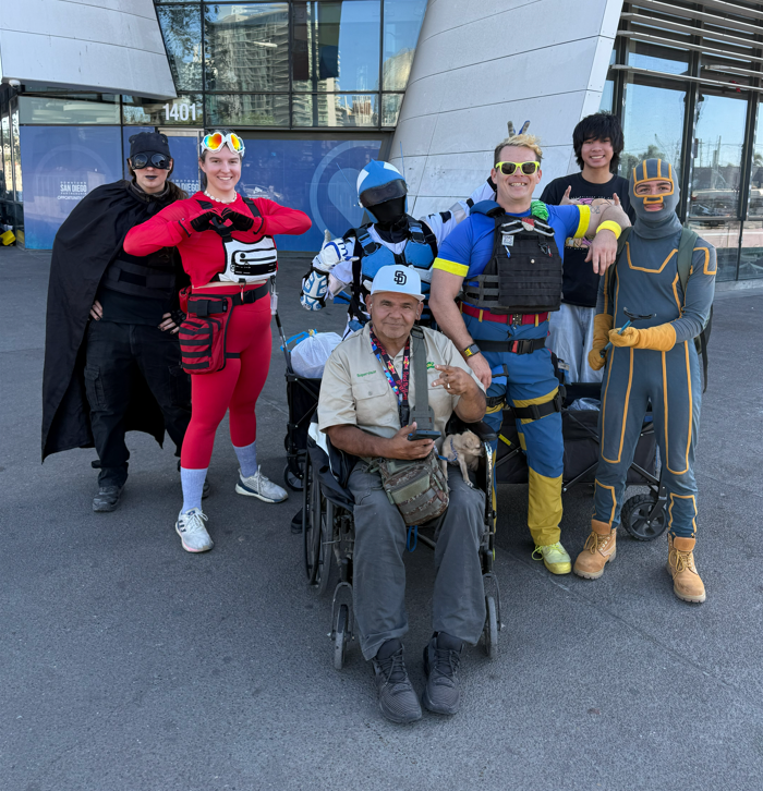 Group of six people in cosplay costumes, one person in a wheelchair, outside a modern building, some making peace signs, with a small dog on the person's lap.