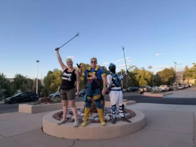 Three people dressed as superheroes in costumes posing on a round platform outdoors, with a parking lot and trees in the background.