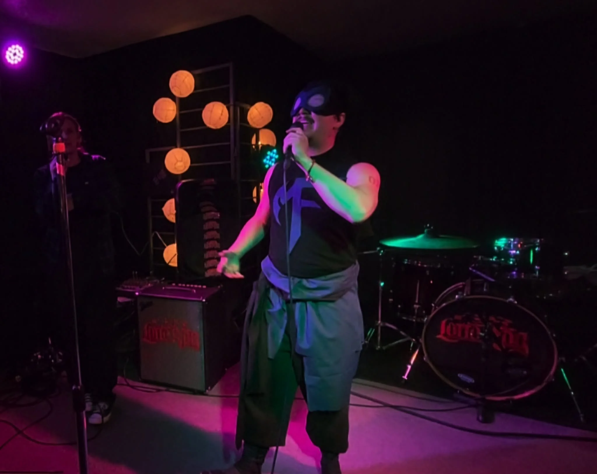 Man with a black mask singing into a microphone at a dark concert with colorful stage lights and lanterns in the background.