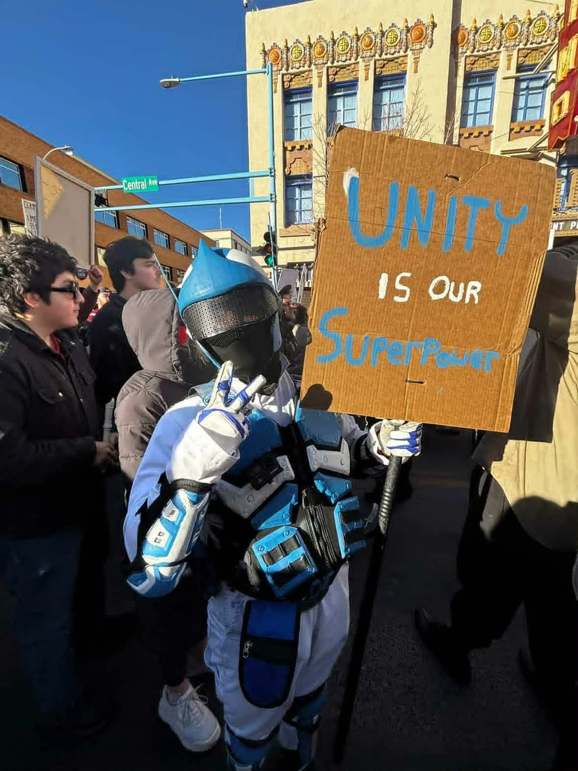 A person in futuristic armor holding a cardboard sign that reads 'Unity is our SuperPower' during a street protest or demonstration, with others in the crowd and city buildings in the background.