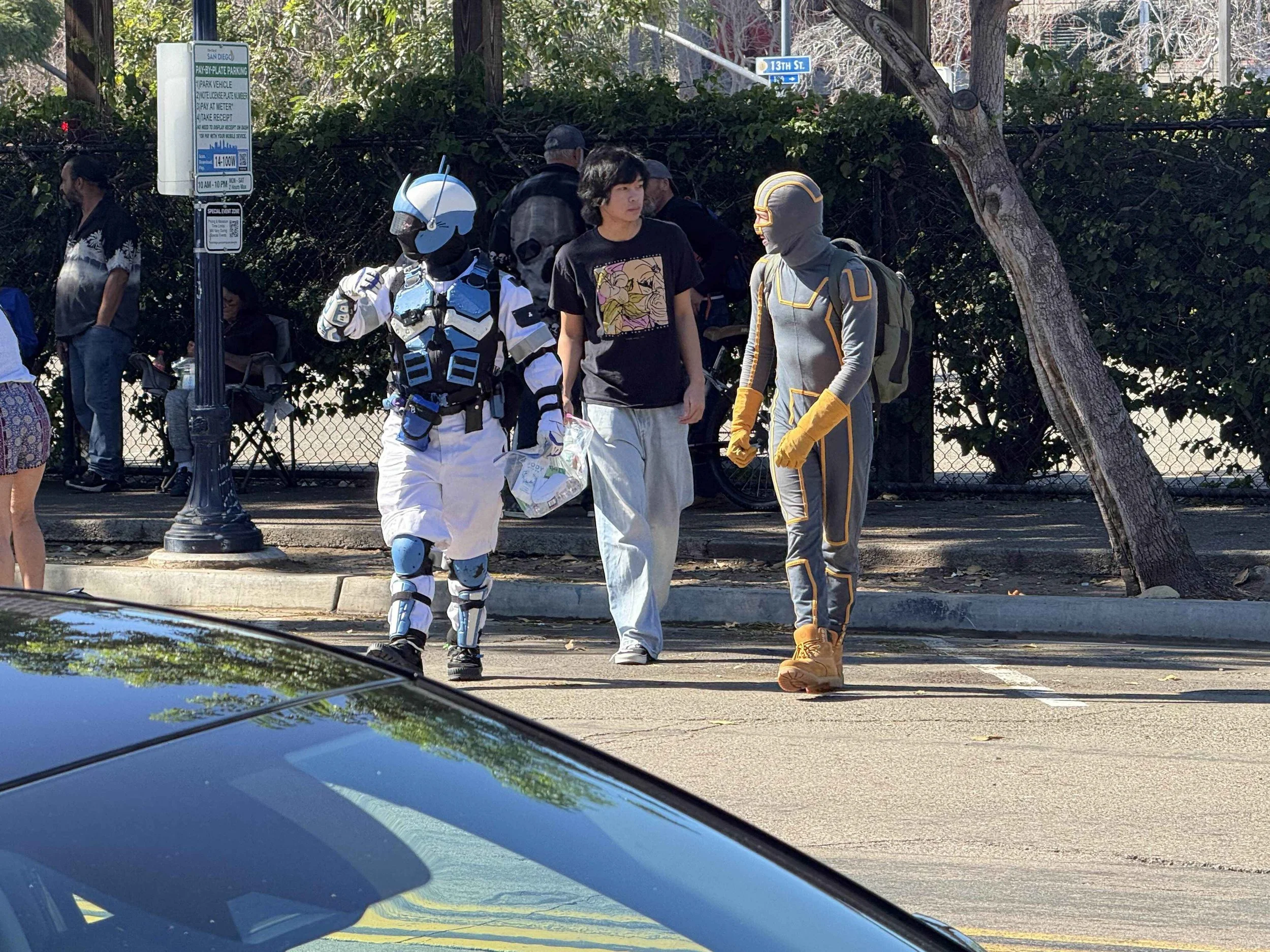 Three people standing on a sidewalk in front of a black fence with trees behind it. Two of them are dressed in sci-fi costumes, one as a sci-fi character with a helmet and armor, and the other in a futuristic suit with gloves and a backpack. The thir