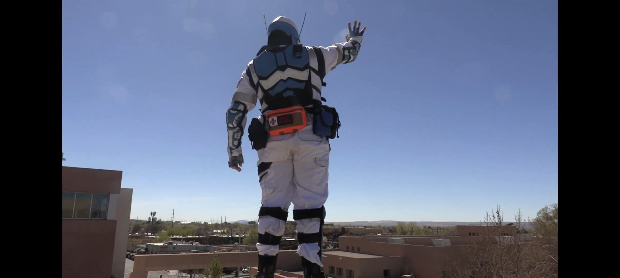 Person dressed in a futuristic or space suit standing on a rooftop, waving at the sky with a clear blue sky and buildings in the background.