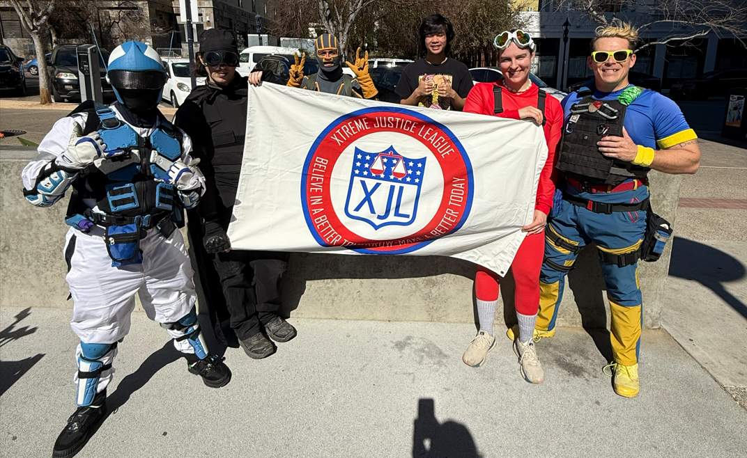Six people dressed in costumes, holding a banner that reads 'XTREME JUSTICE LEAGUE' with a logo in the center, standing outdoors on a sidewalk.