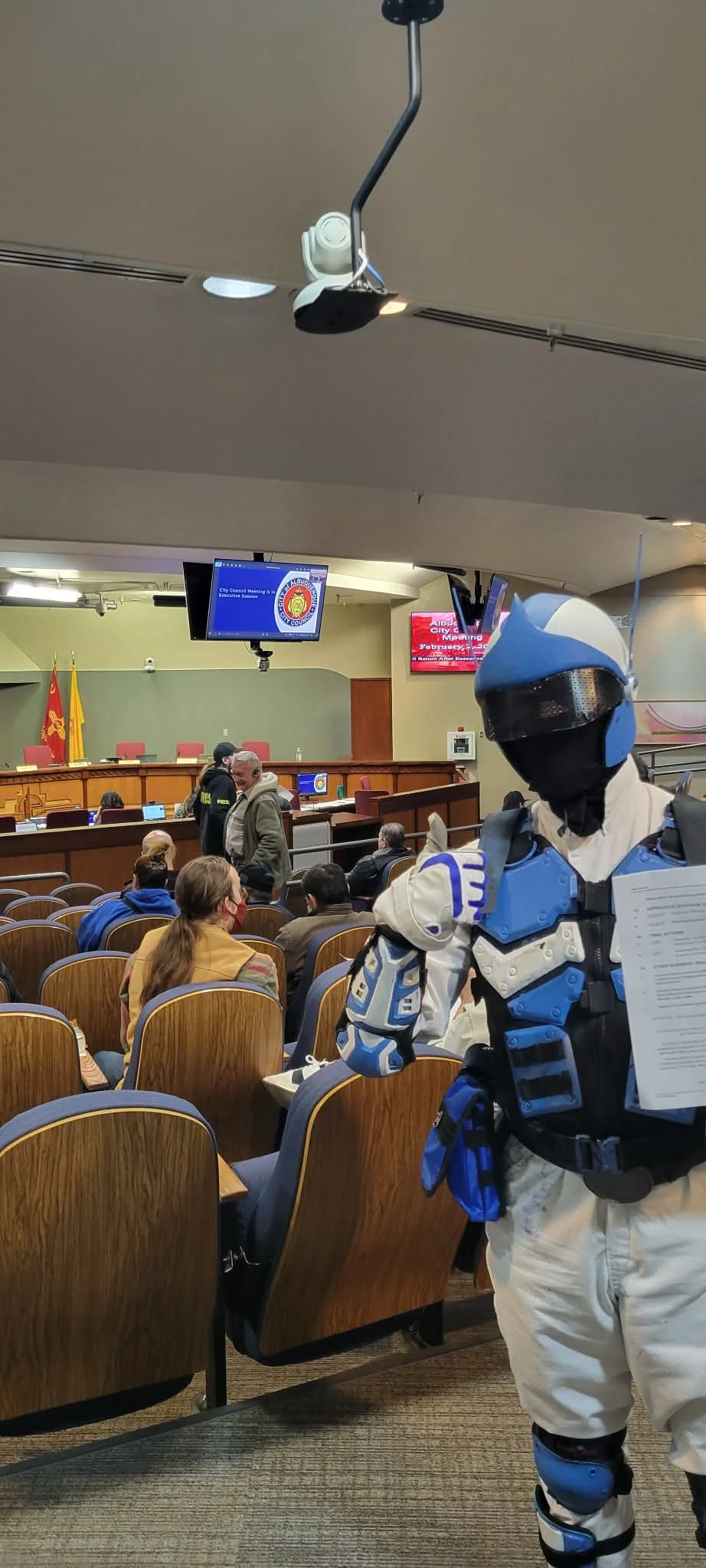 A person dressed in a futuristic blue and white robot costume holding a paper in a government meeting room with people seated and a large screen displaying a city council logo in the background.