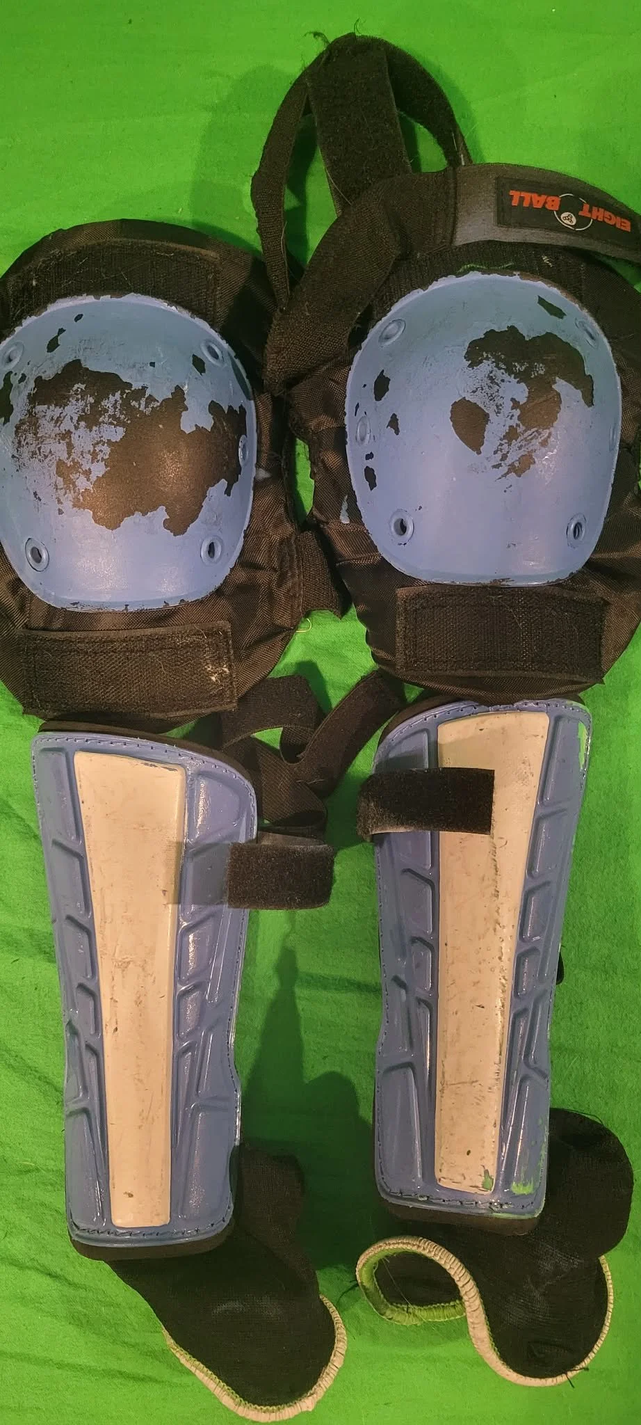 A pair of baseball catcher's leg guards and knee guards with black straps and padding, placed on a green surface.