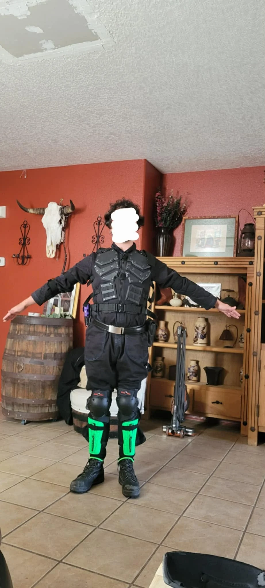 Person dressed in tactical or combat gear, standing indoors with arms outstretched, in front of a wooden shelf with vases, framed pictures, and a mounted animal skull with horns on a red wall.