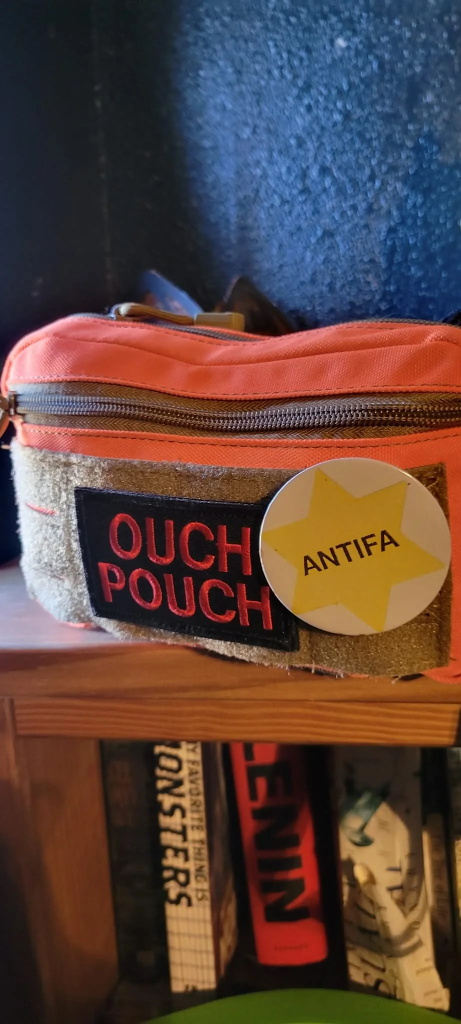 A small pouch with a patch saying 'OUCH POUCH' and a sticker that reads 'ANTIFA' with a yellow star. The pouch appears to be made of fabric and leather, with a zipper on top.