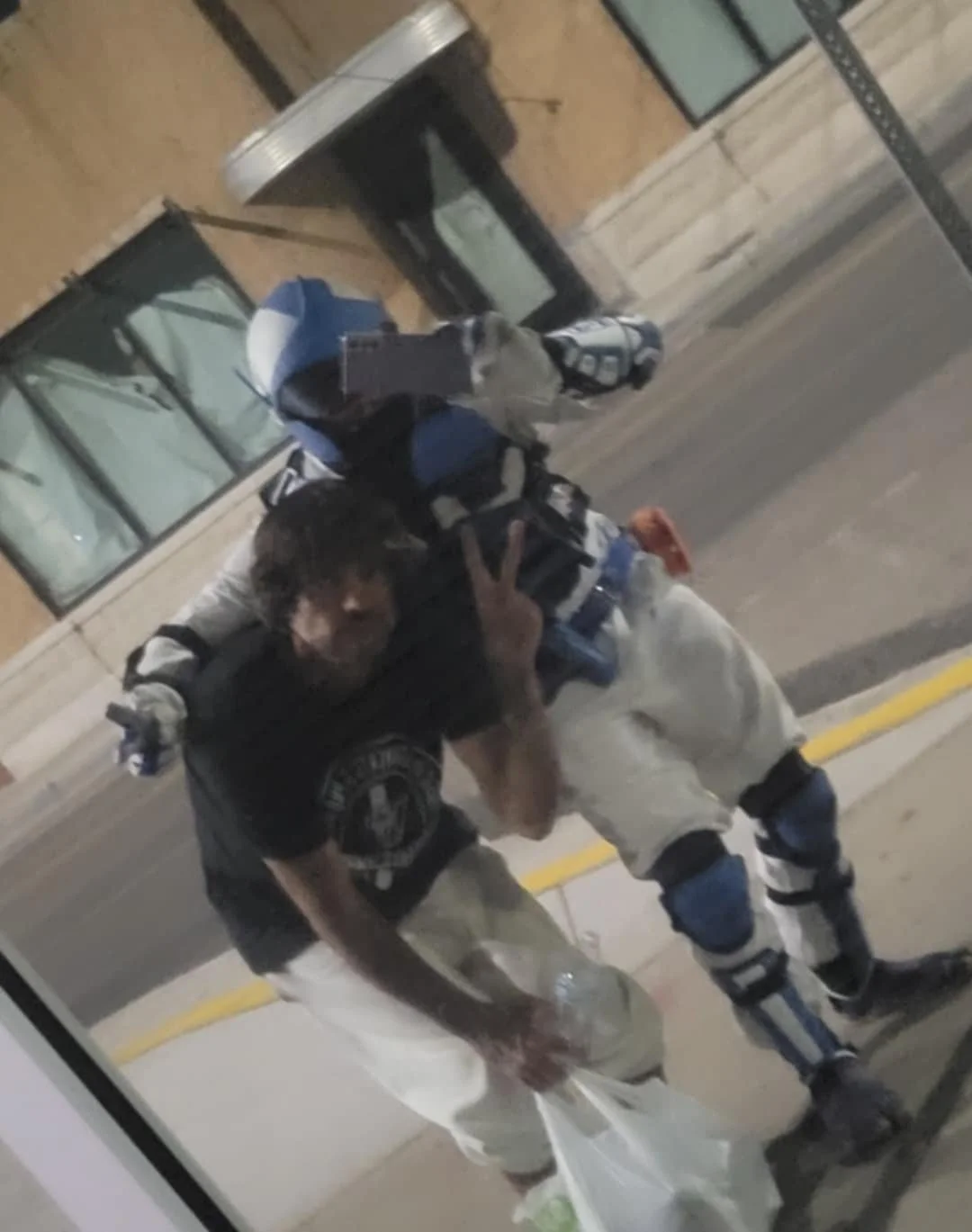 A young man with dark hair taking a mirror selfie while making a peace sign with his right hand. He is wearing a black T-shirt with a circular design and white pants with protective knee guards. He is carrying a blue and black backpack and has a whit