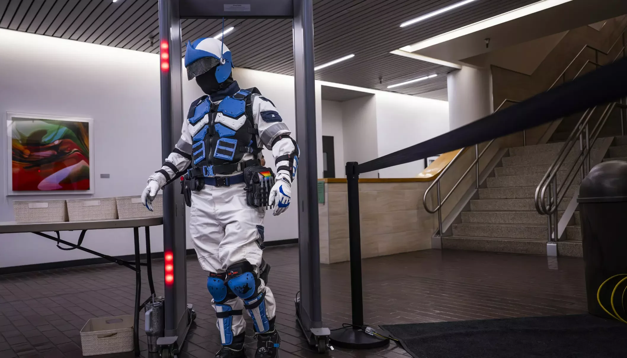 Person wearing a full-body futuristic, high-tech suit with blue and white armor-like padding, helmet, and gloves, walking through a security checkpoint with a center scanning device in an indoor public space. The scene includes a staircase, artwork o