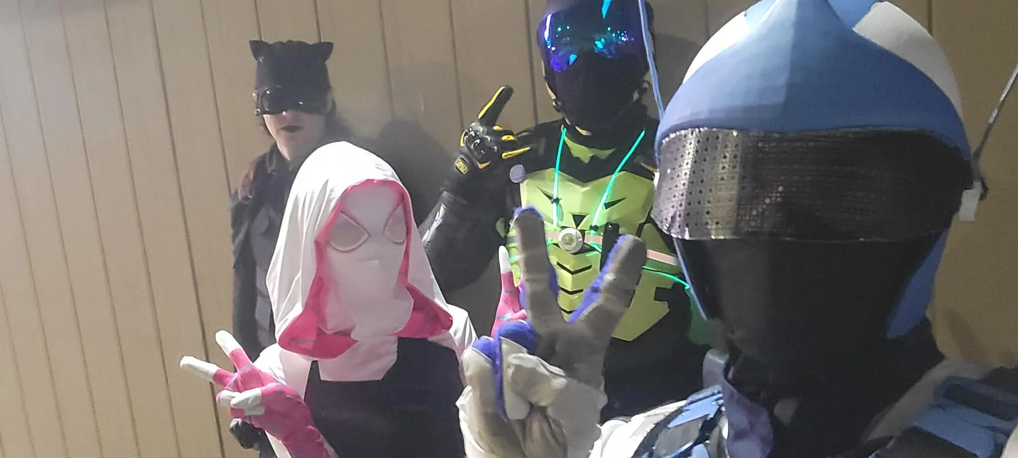 Four individuals wearing superhero or costume cosplay gear posing for a photo indoors against a wooden-paneled wall.