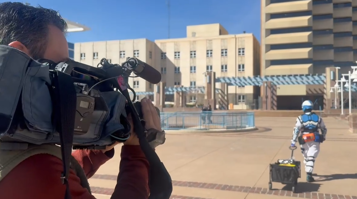 A cameraman filming a person dressed as a futuristic soldier in a blue and white suit with an orange toolbox, in an outdoor urban area with buildings and a clear blue sky.