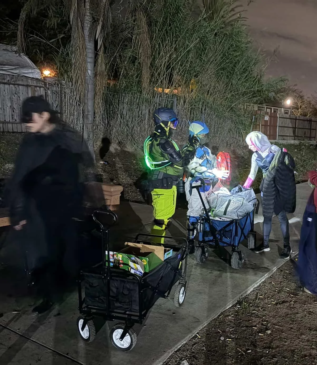 Group of people, including emergency responders, gathered outdoors at night sorting through supplies near a fence and trees.