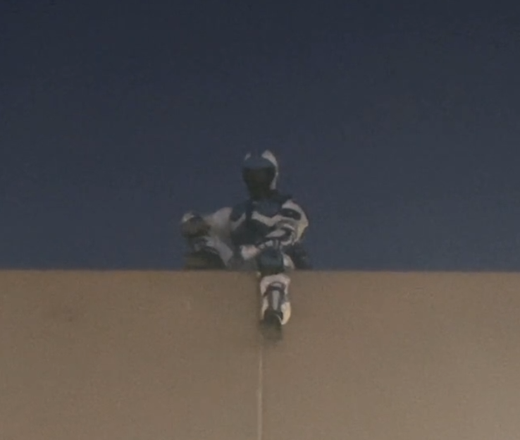A person dressed in a racing suit and helmet, sitting on the edge of a roof or ledge, looking down.