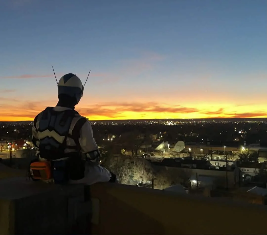 Person wearing a helmet and emergency vest sitting on a ledge watching sunset over city with radio dishes and buildings below.