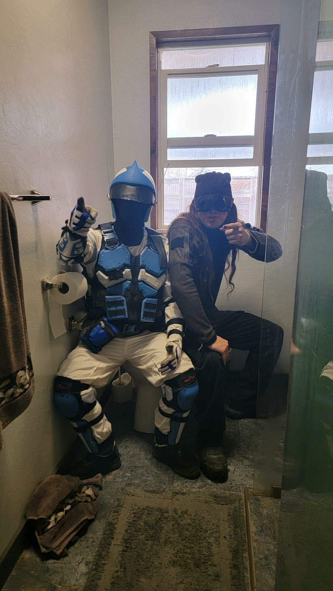 Two people dressed in cosplay costumes seated in a small bathroom, taking a mirror selfie. One is in a blue and black armor suit resembling a sci-fi or futuristic character, giving a thumbs-up. The other is in casual attire with a headband equipped w