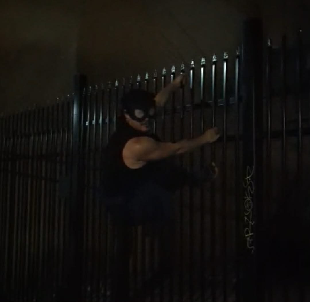 A person dressed as a criminal wearing a bunny mask and holding a crowbar, attempting to climb a tall metal fence at night.