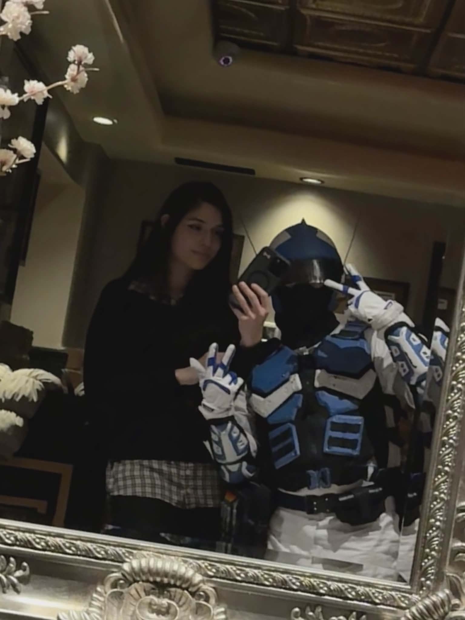 A woman taking a selfie in a mirror along with a person dressed as a robot in a room with decorative elements and wooden furniture.