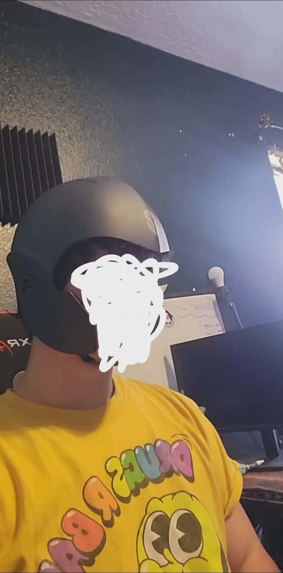 Person wearing a yellow T-shirt with a colorful cartoon graphic, black cycling helmet, and face mask, sitting in a room with computer monitors and electronic equipment.