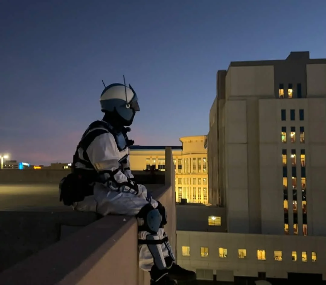 Person dressed in a robot or futuristic costume sitting on a ledge on a rooftop, overlooking the city buildings at dusk.