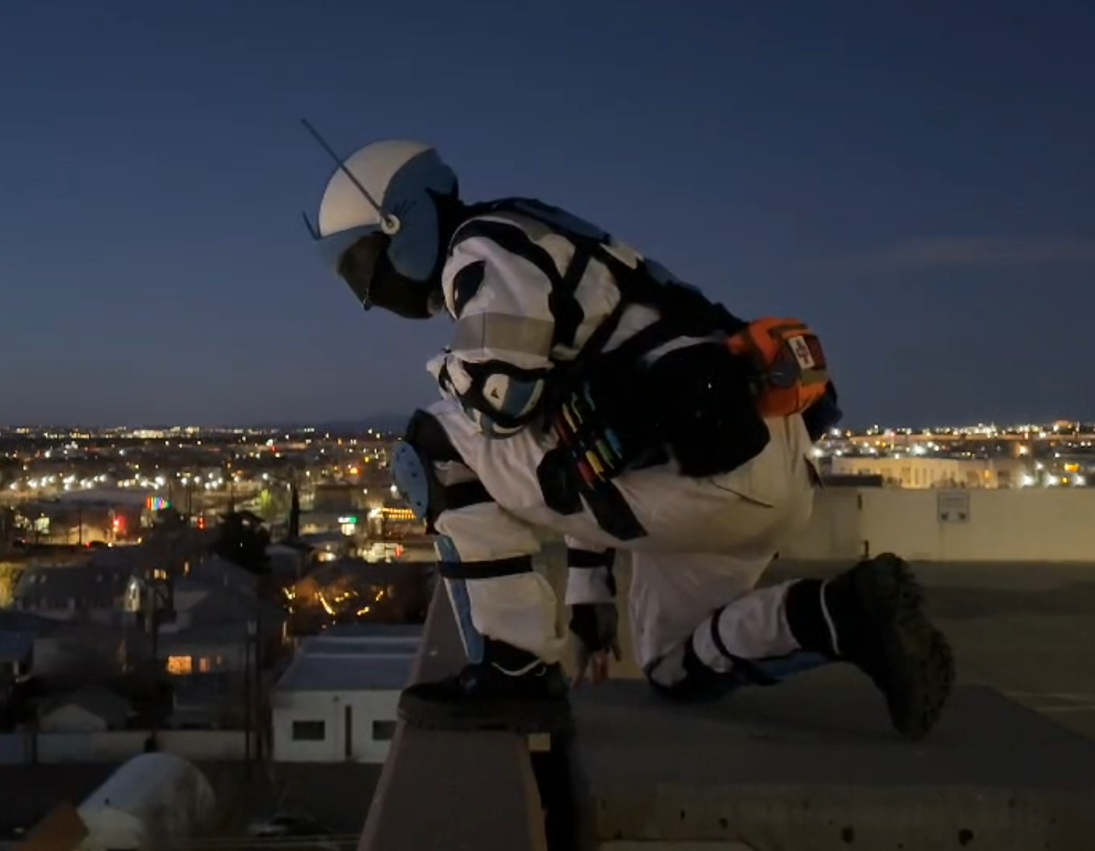 A rescue worker kneeling on a rooftop at dusk, wearing a white and black uniform with a helmet, harness, and safety equipment, overlooking a cityscape with lights in the distance.