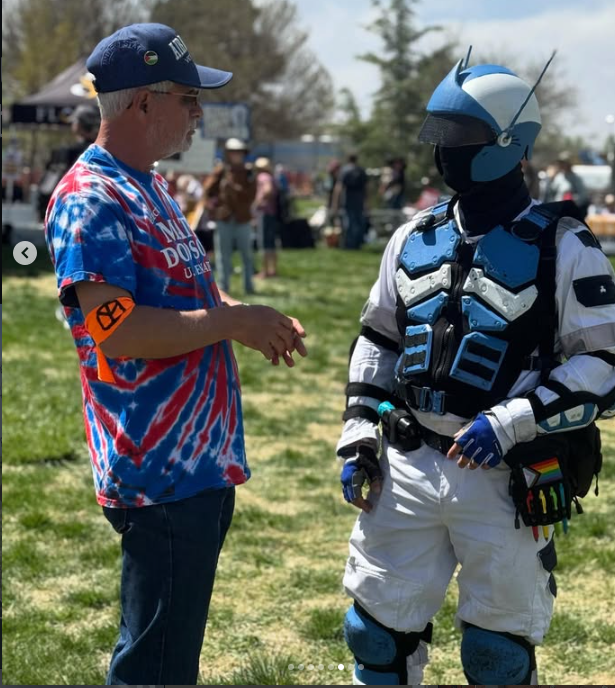 A man in a blue tie-dye shirt and a navy blue cap talking to a person dressed in futuristic blue and white armor with a helmet and vest, in an outdoor setting with grass and trees, during a daytime event.