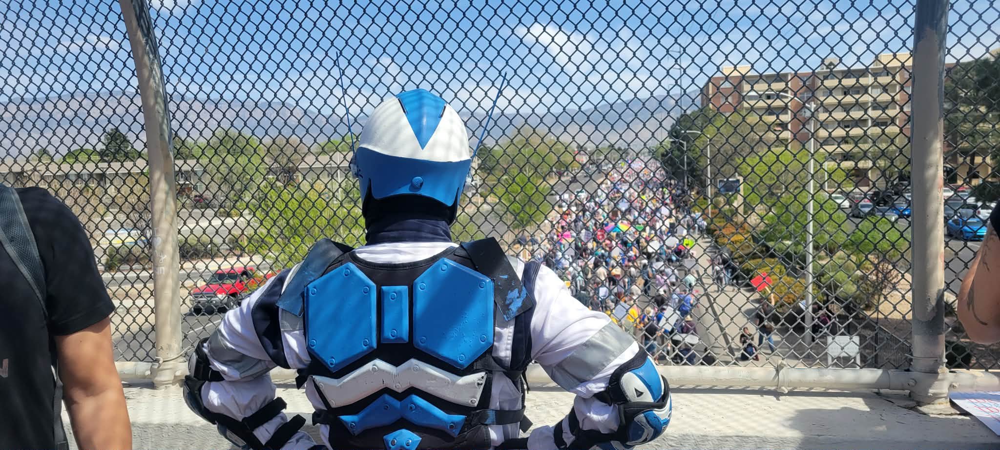 A person dressed in a blue and white costume, possibly a superhero or futuristic warrior, standing behind a chain-link fence and looking down at a large crowd of people walking along a street, with buildings and trees in the background.