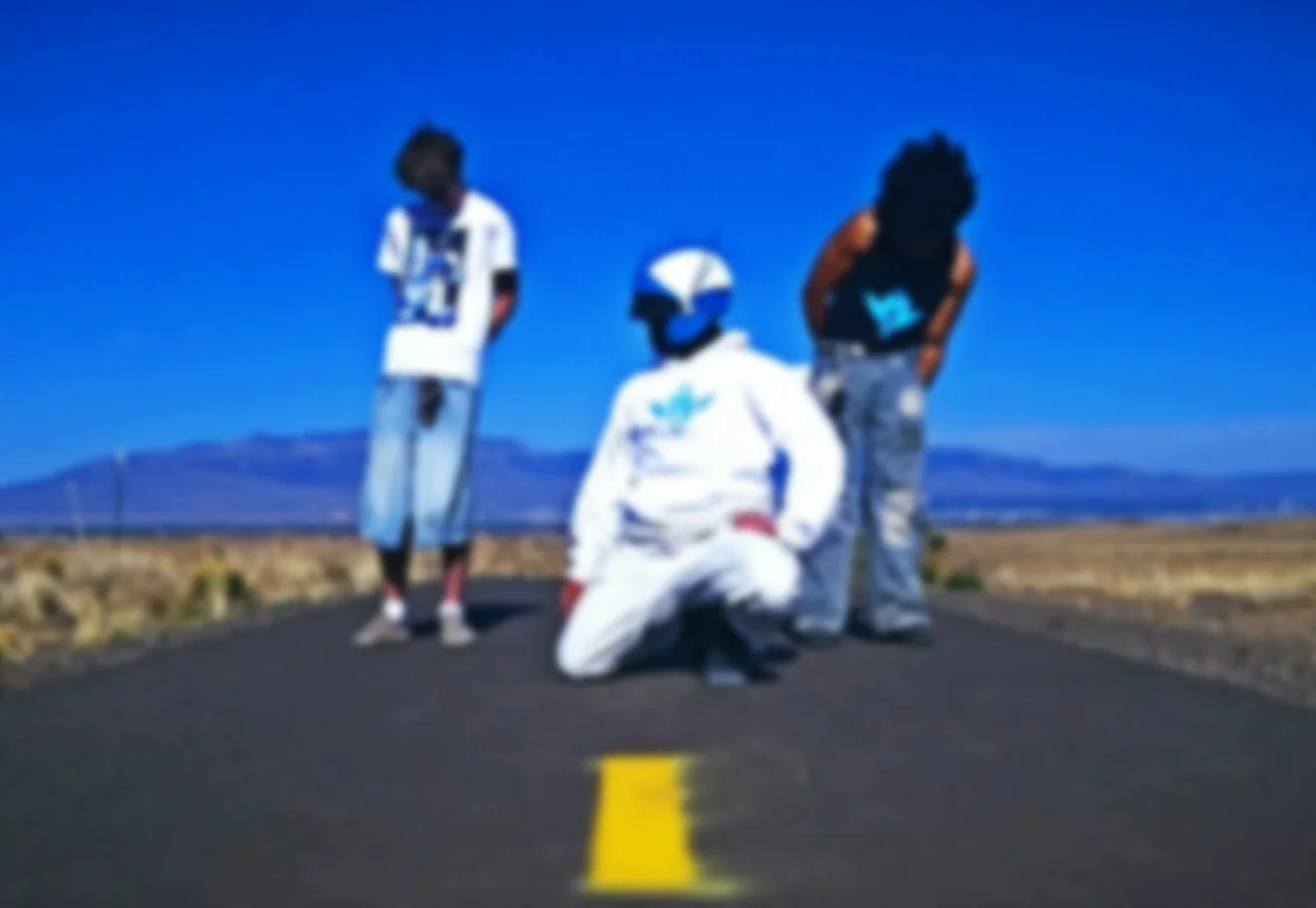 Three people on a deserted road in a wide-open landscape with mountains in the background, under a clear blue sky. One person is kneeling in front wearing a racing helmet and suit, while the other two stand nearby, looking towards the camera.